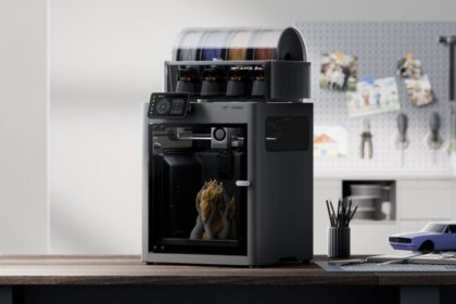 Desktop 3D printer on a wooden workbench in a workshop, with spools of filament on top and tools on a pegboard in the background.