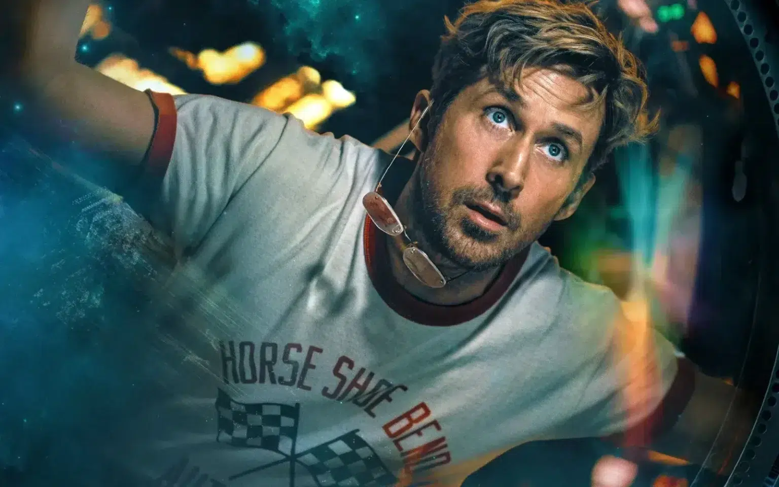 Man with stubble peers through a circular porthole, wearing a light gray T-shirt and sunglasses hanging from his neck inside a vehicle or capsule.