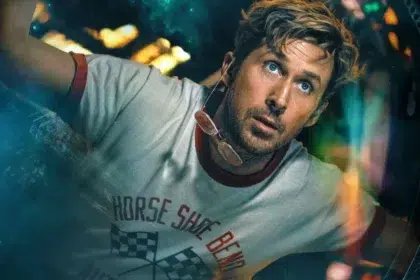 Man with stubble peers through a circular porthole, wearing a light gray T-shirt and sunglasses hanging from his neck inside a vehicle or capsule.