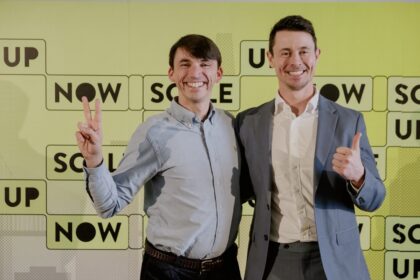Two men smiling at camera; one shows peace sign, the other thumbs up, in front of a bright yellow backdrop with blocks that say NOW and UP.