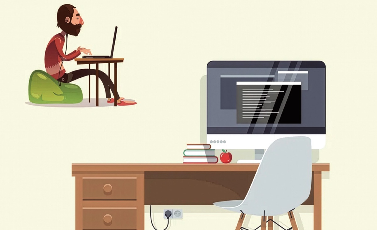 Cartoon of a bearded person sitting on a green bean bag and typing on a laptop at a desk with a desktop computer, books, and an apple nearby.