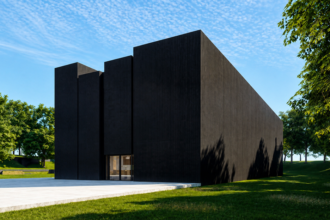 Modern black brick building with tall, vertical walls and a glass entrance set in a green park under a blue sky with scattered clouds