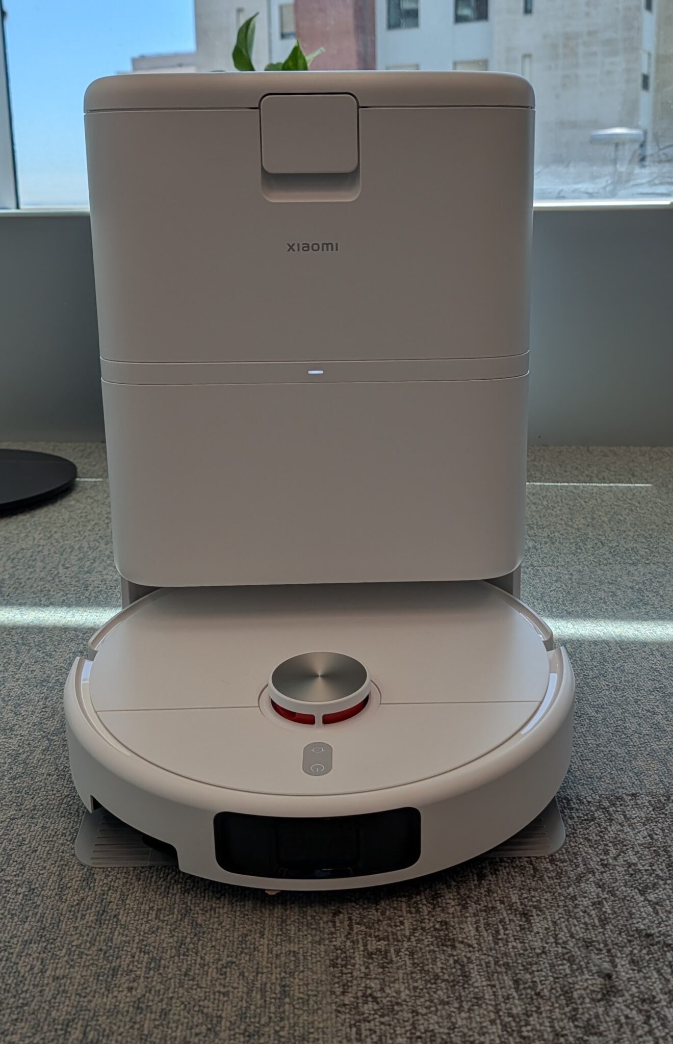 White Xiaomi robotic vacuum with its charging dock behind it, lid open, indoors near a window.