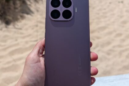 Purple iPhone held in a hand, showing a square quad-camera module against a sandy beach background, rear view.