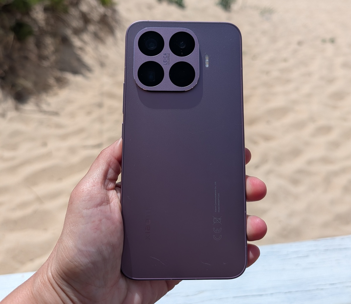 Purple iPhone held in a hand, showing a square quad-camera module against a sandy beach background, rear view.