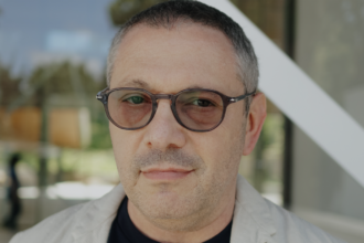 Close-up portrait of a man with gray-stubbled beard and brown round glasses, outdoors near a glass building.