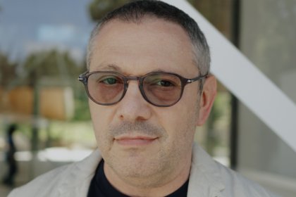 Close-up portrait of a man with gray-stubbled beard and brown round glasses, outdoors near a glass building.