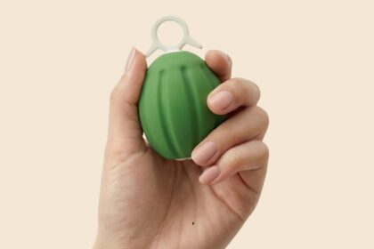 Hand holding a small green grenade-shaped plastic fidget toy with a white ring on top.