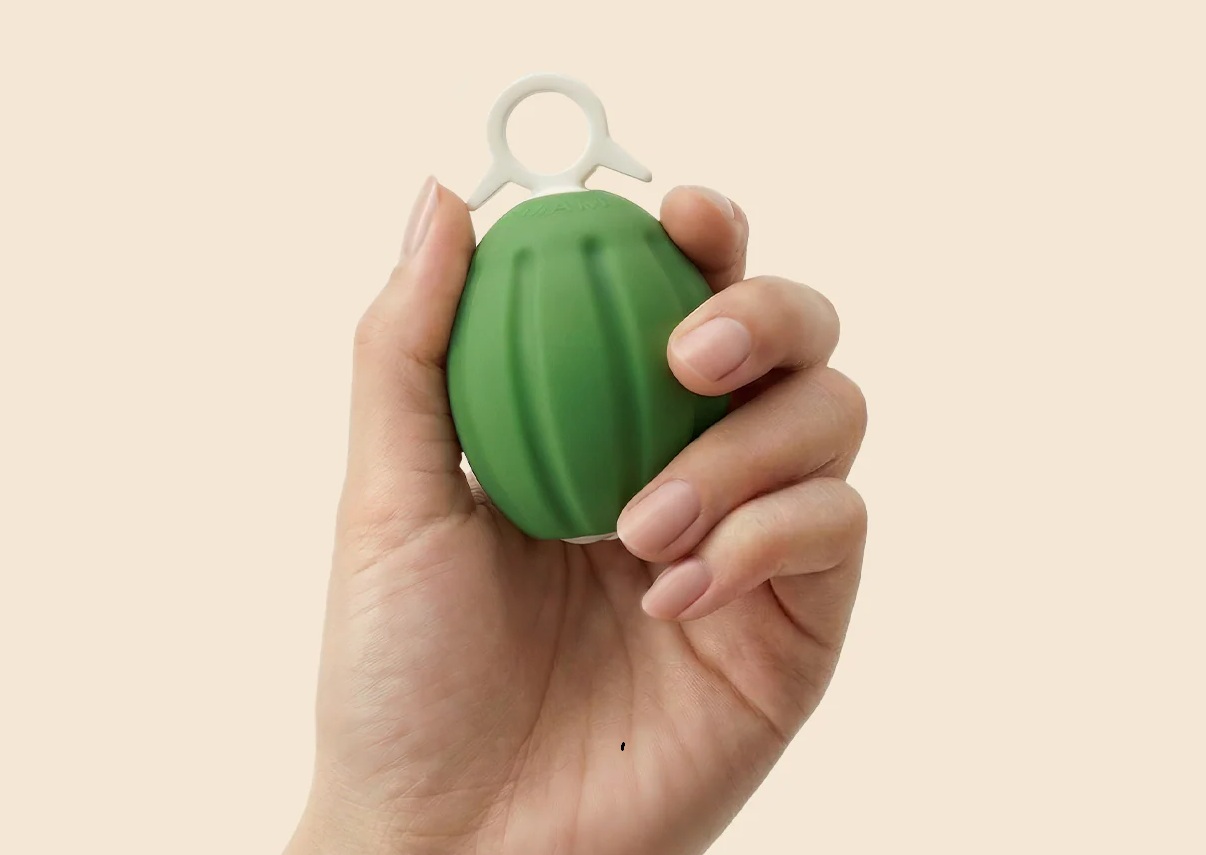 Hand holding a small green grenade-shaped plastic fidget toy with a white ring on top.