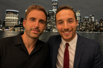 Two men pose for a photo at night by a harbor, one in a black polo and the other in a suit and red tie, city lights behind them.