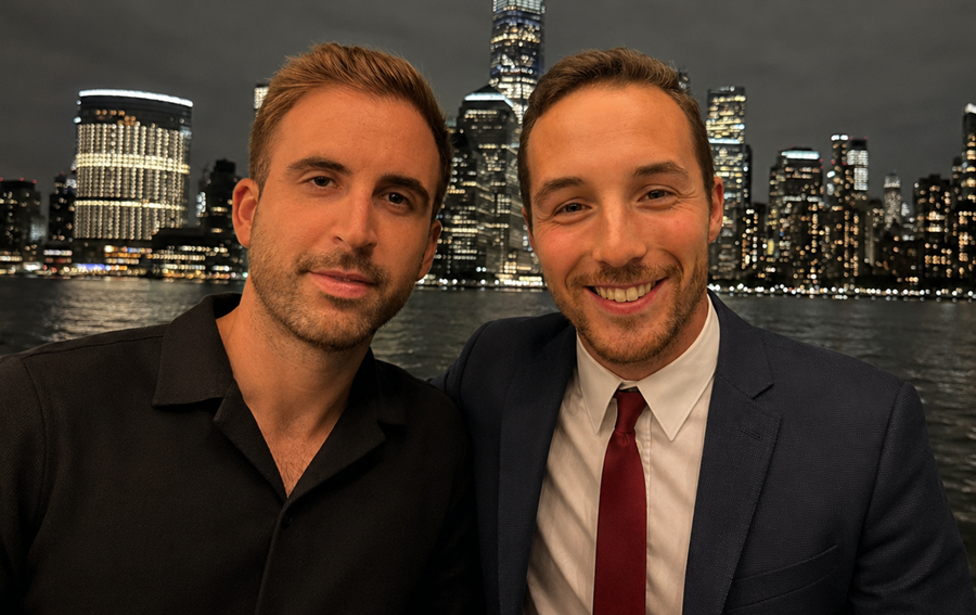 Two men pose for a photo at night by a harbor, one in a black polo and the other in a suit and red tie, city lights behind them.