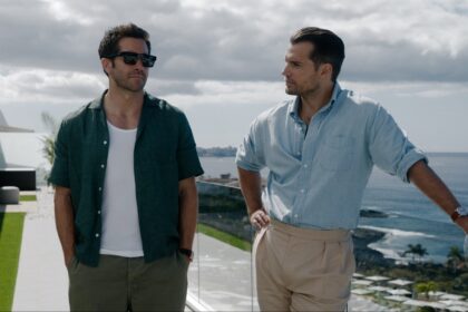 Two men stand on a sunny coastal balcony, chatting by a glass railing with the sea and sky in the background. One wears a green shirt and sunglasses, the other a light blue shirt and beige pants.