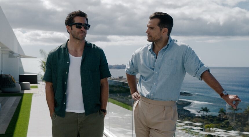 Two men stand on a sunny coastal balcony, chatting by a glass railing with the sea and sky in the background. One wears a green shirt and sunglasses, the other a light blue shirt and beige pants.