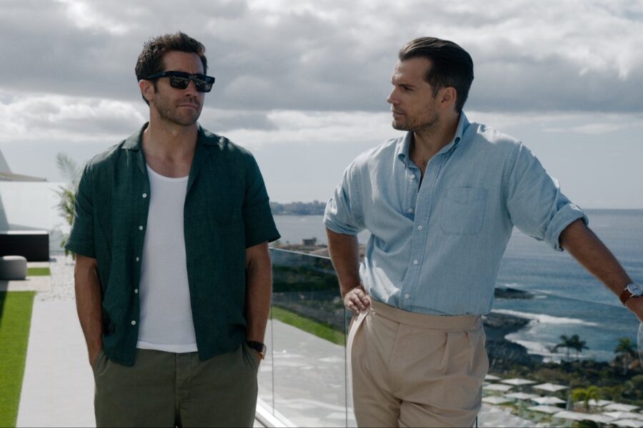 Two men stand on a sunny coastal balcony, chatting by a glass railing with the sea and sky in the background. One wears a green shirt and sunglasses, the other a light blue shirt and beige pants.