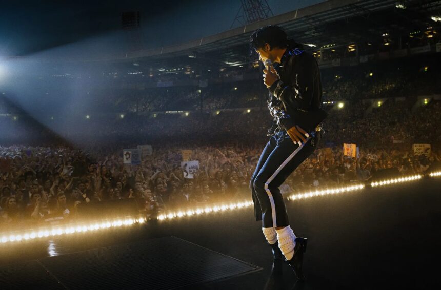 Performer on stage singing into a microphone to a massive crowded arena at night, bright stage lights behind him.