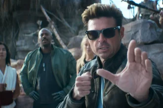 Man in sunglasses and a brown jacket raises a defensive-hand gesture in the foreground while a small group stands behind him in a rocky outdoor setting.