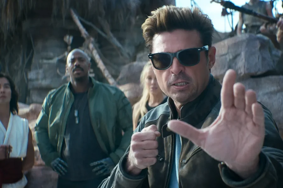 Man in sunglasses and a brown jacket raises a defensive-hand gesture in the foreground while a small group stands behind him in a rocky outdoor setting.