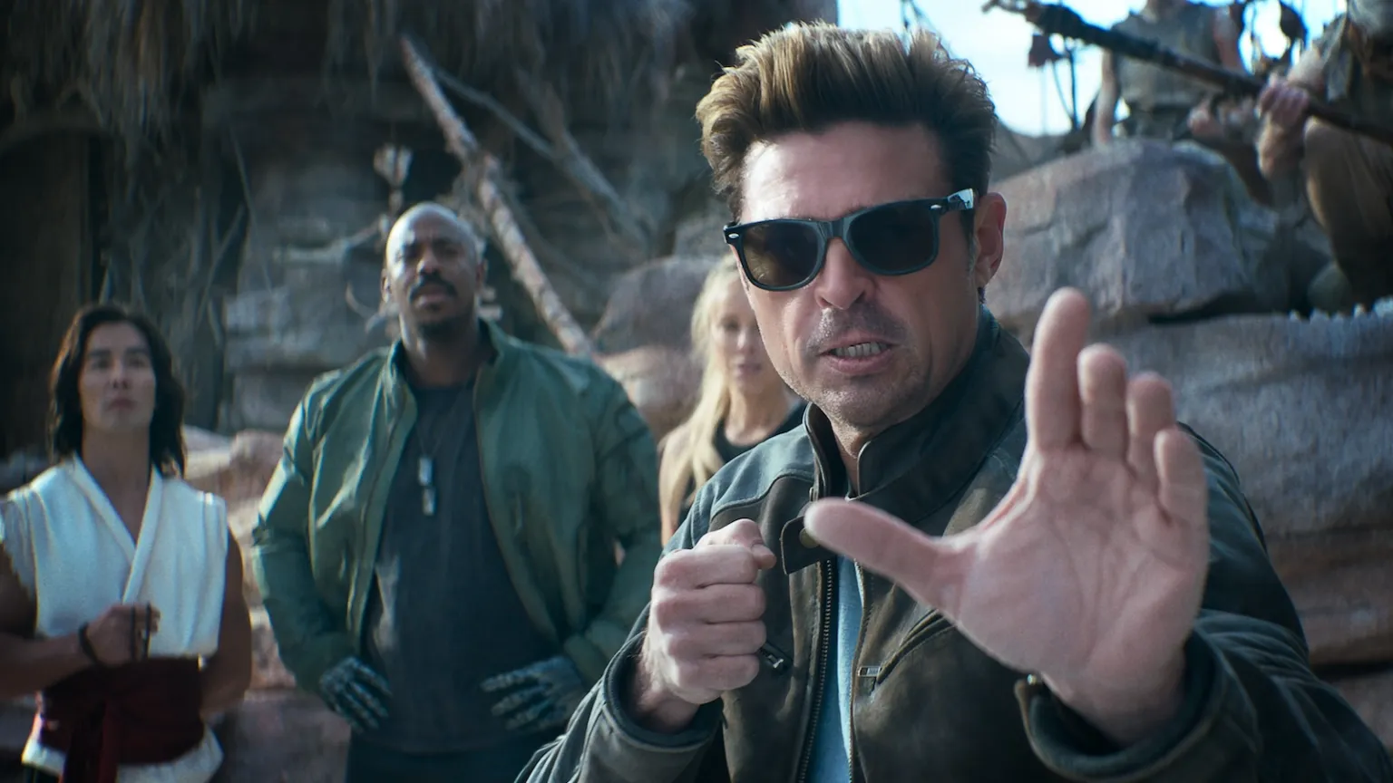 Man in sunglasses and a brown jacket raises a defensive-hand gesture in the foreground while a small group stands behind him in a rocky outdoor setting.