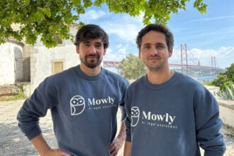Two men in matching navy Mowly sweatshirts pose outdoors under a leafy tree, with a red suspension bridge in the background.