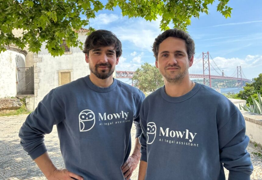 Two men in matching navy Mowly sweatshirts pose outdoors under a leafy tree, with a red suspension bridge in the background.