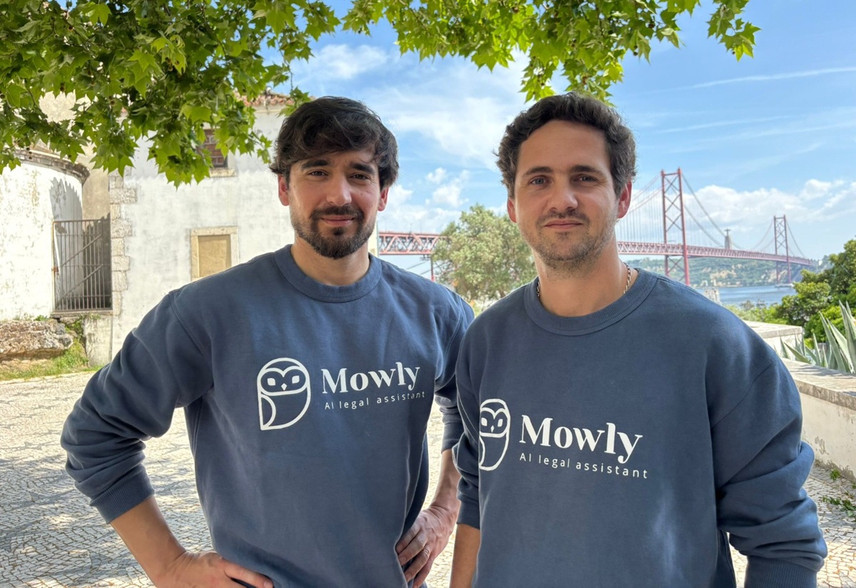 Two men in matching navy Mowly sweatshirts pose outdoors under a leafy tree, with a red suspension bridge in the background.