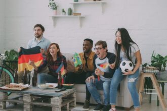 Group of diverse friends cheering in a bright living room while watching a soccer game on the TV, waving Germany and Brazil flags with pizza and drinks on a rustic coffee table nearby.
