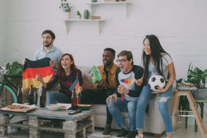 Group of diverse friends cheering in a bright living room while watching a soccer game on the TV, waving Germany and Brazil flags with pizza and drinks on a rustic coffee table nearby.