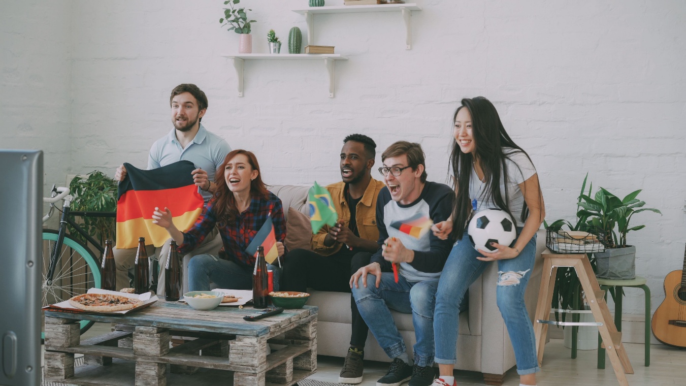 Group of diverse friends cheering in a bright living room while watching a soccer game on the TV, waving Germany and Brazil flags with pizza and drinks on a rustic coffee table nearby.