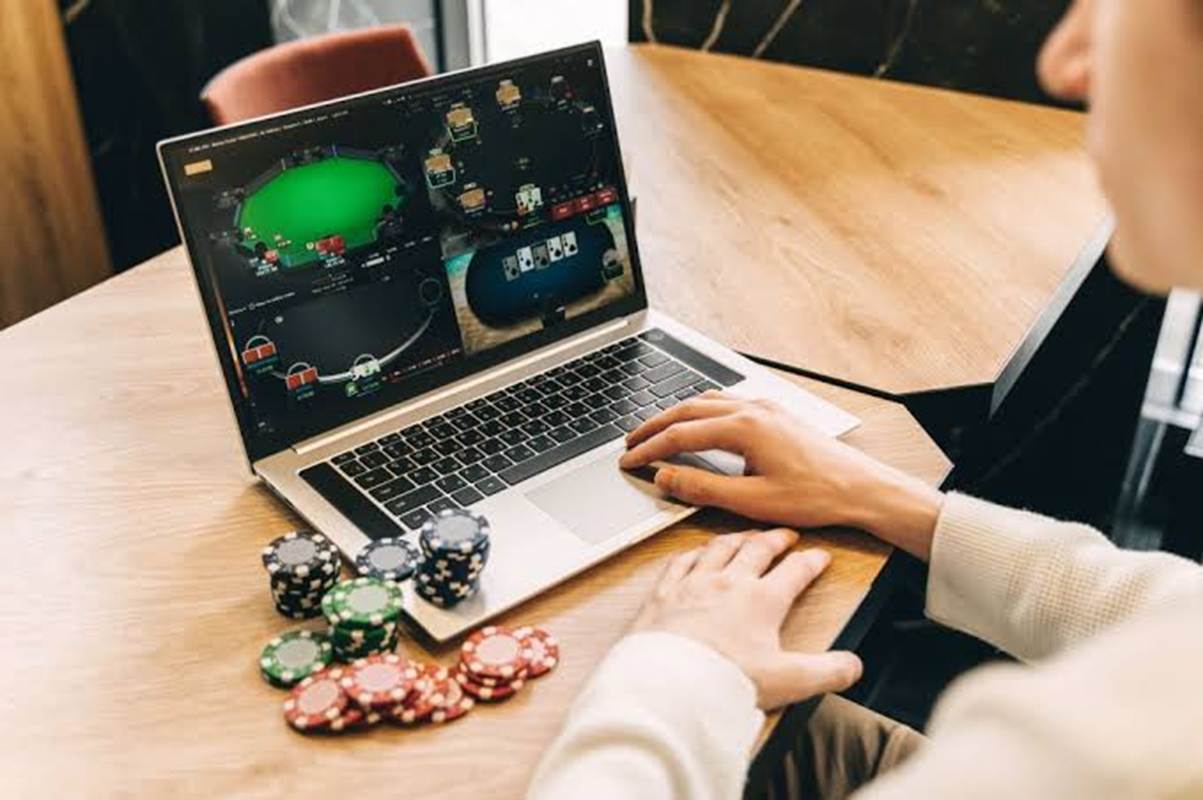 Person at a wooden table using a laptop to play online poker, with stacked poker chips beside the computer.