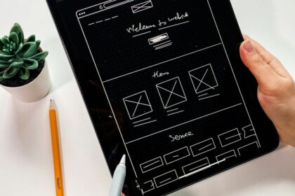 Hands hold a tablet showing a rough website wireframe sketch, with a pencil and succulent on a white desk nearby.