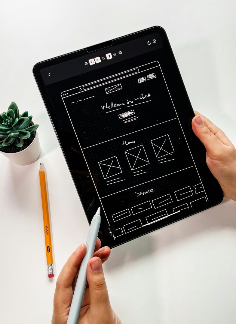 Hands hold a tablet showing a rough website wireframe sketch, with a pencil and succulent on a white desk nearby.