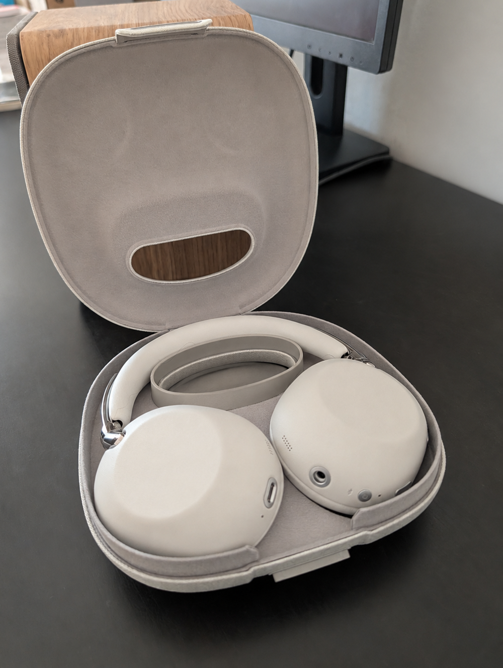 Light gray over-ear headphones resting in an open soft carrying case on a dark surface, showing the case interior.