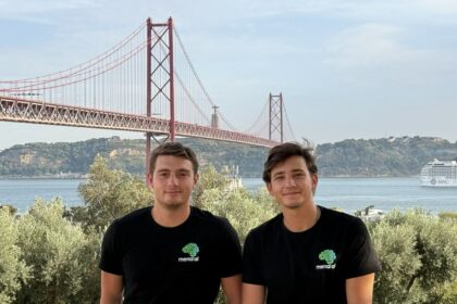 Two men in black shirts with a green brain logo pose in front of a red suspension bridge over water, with trees and a cruise ship in the background.