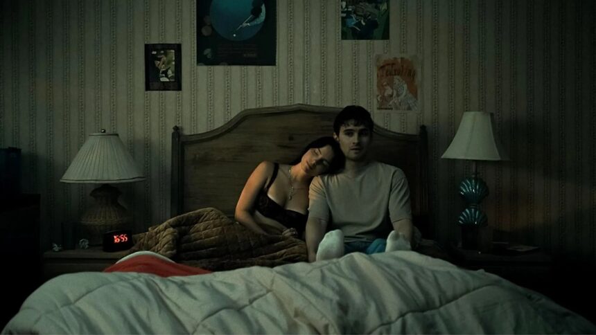 Dimly lit bedroom scene with a couple sitting up in bed under a quilt; woman rests her head on the man's shoulder amid vintage wallpaper and lamps on each side.