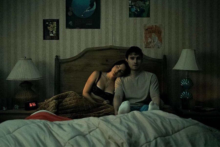 Dimly lit bedroom scene with a couple sitting up in bed under a quilt; woman rests her head on the man's shoulder amid vintage wallpaper and lamps on each side.
