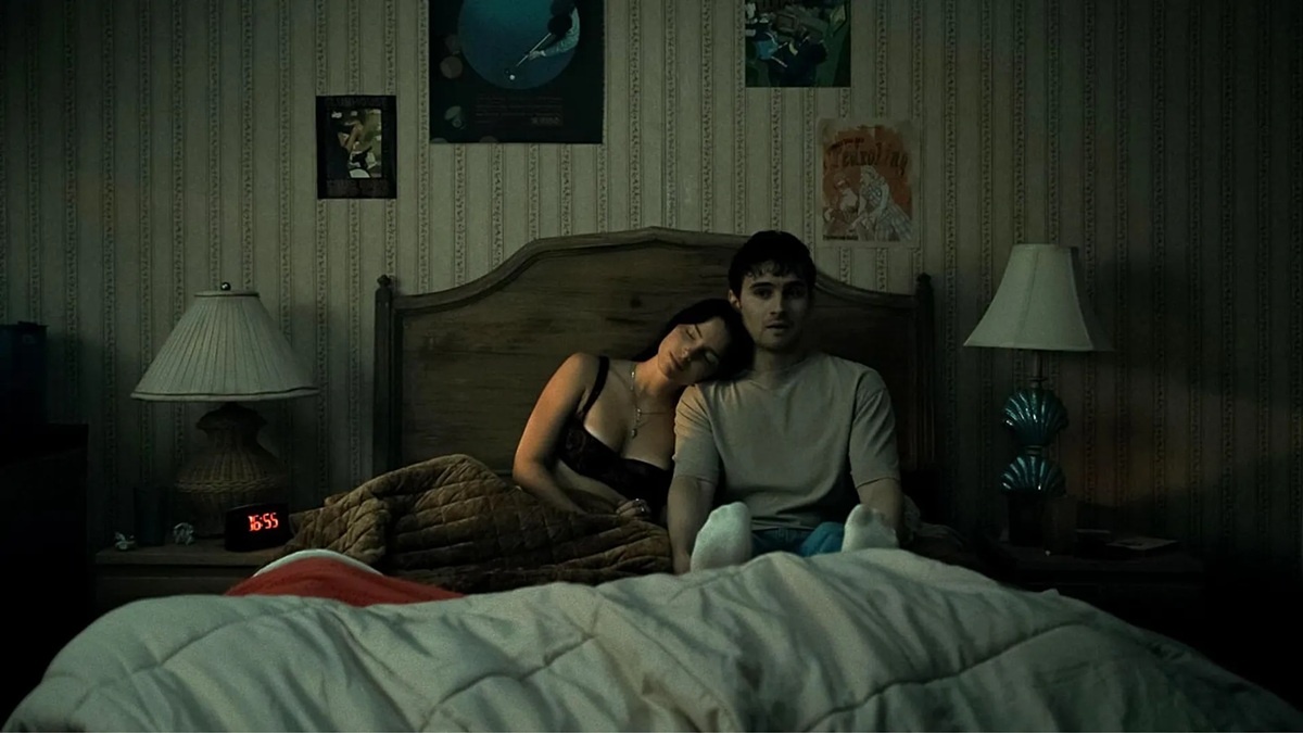 Dimly lit bedroom scene with a couple sitting up in bed under a quilt; woman rests her head on the man's shoulder amid vintage wallpaper and lamps on each side.