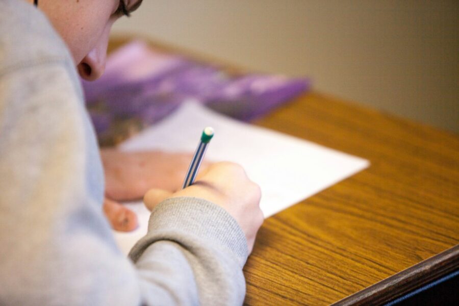 Close-up of a person’s hand writing with a striped blue pencil on a white sheet at a wooden desk.