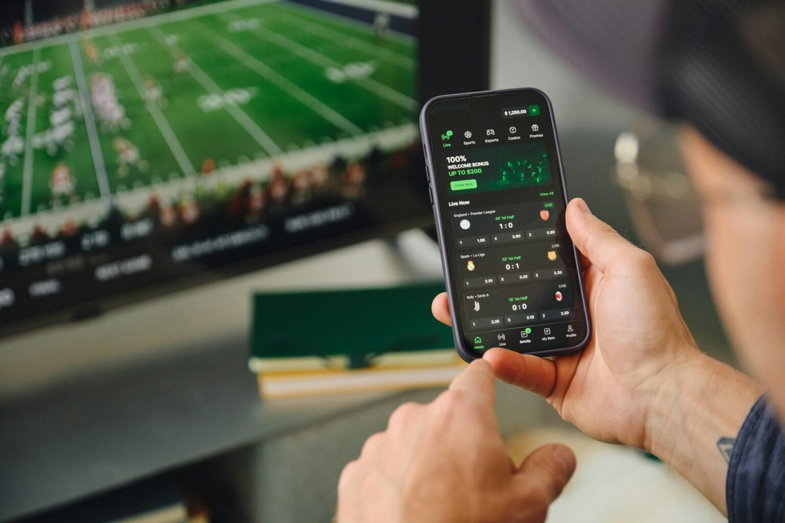Hand holding a smartphone displaying a sports betting app while a football match plays on a blurred TV screen in the background.