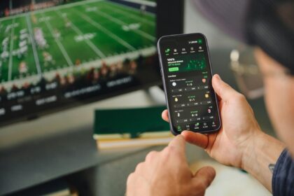 Hand holding a smartphone displaying a sports betting app while a football match plays on a blurred TV screen in the background.