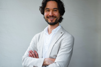 Smiling man with dark hair and beard, wearing a light blazer and white shirt, arms crossed against a neutral background.