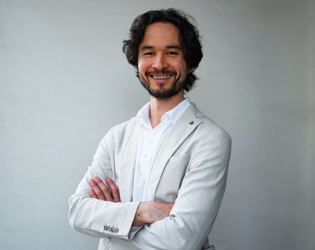 Smiling man with dark hair and beard, wearing a light blazer and white shirt, arms crossed against a neutral background.