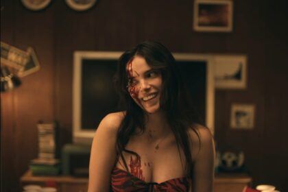 Woman wearing a red and black striped top with blood makeup on her face and chest, smiling in a warmly lit, retro living room with a TV behind her.