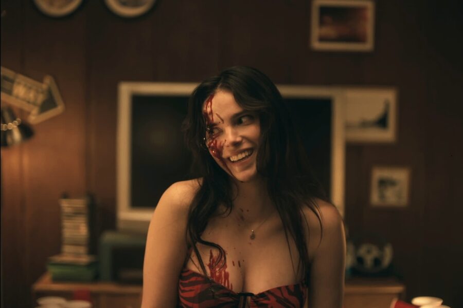 Woman wearing a red and black striped top with blood makeup on her face and chest, smiling in a warmly lit, retro living room with a TV behind her.