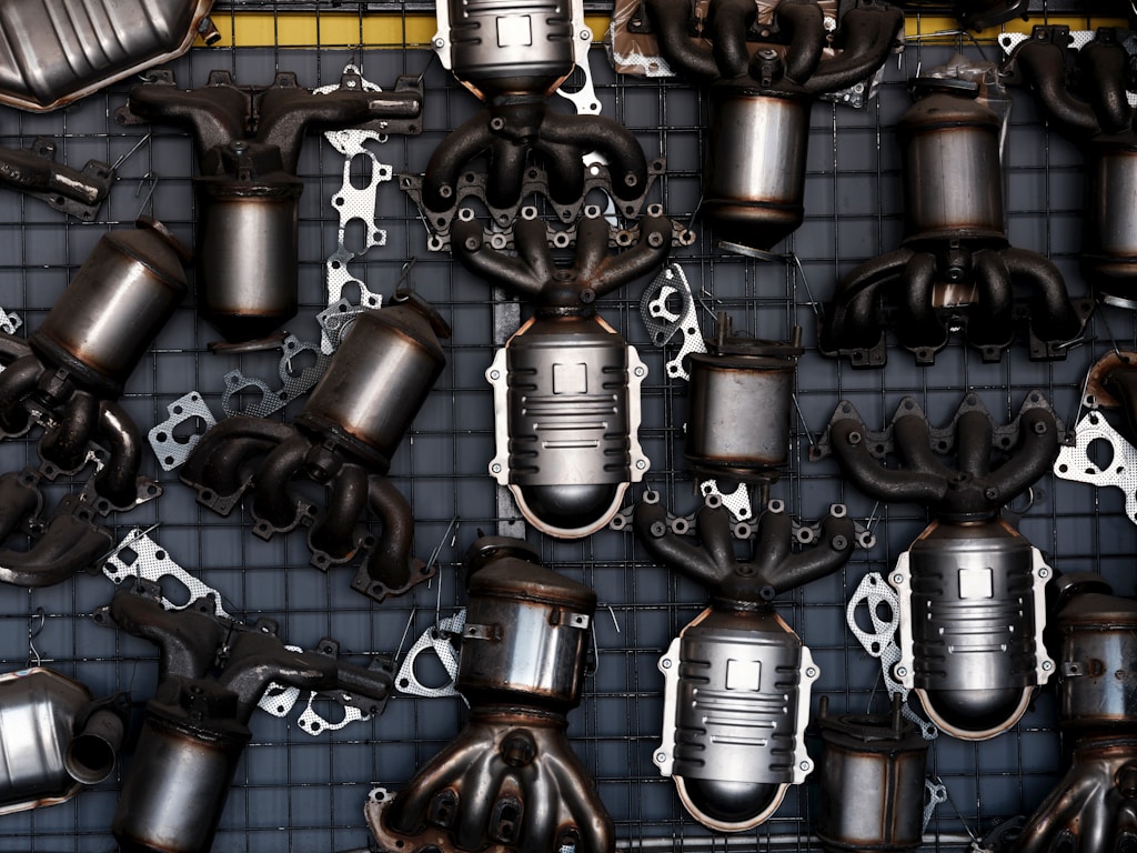 A dense arrangement of car exhaust parts, including catalytic converters and exhaust manifolds, laid out on a metal grid.