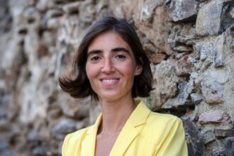 Woman with short brown hair in a yellow blazer leaning against a stone wall, smiling at the camera.