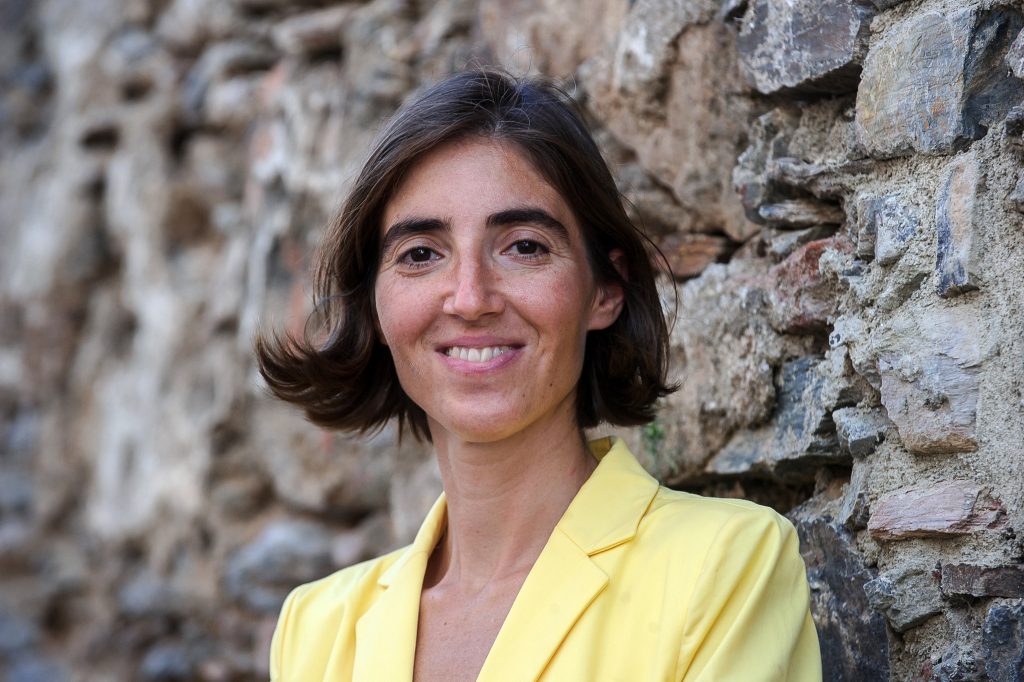 Woman with short brown hair in a yellow blazer leaning against a stone wall, smiling at the camera.