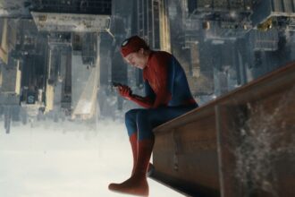 Spider-Man in red-blue suit sits on a rooftop ledge, looking at a smartphone above a vast cityscape below the skyline.
