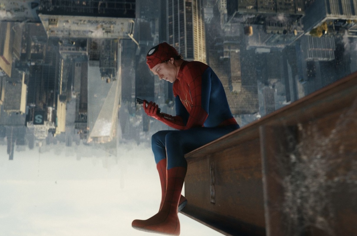 Spider-Man in red-blue suit sits on a rooftop ledge, looking at a smartphone above a vast cityscape below the skyline.
