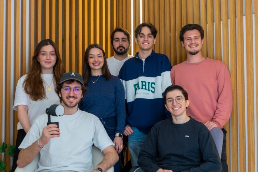 Group of seven young adults posing for a photo in front of a vertical wooden slat wall; front-left person sits holding a handheld microphone strap.
