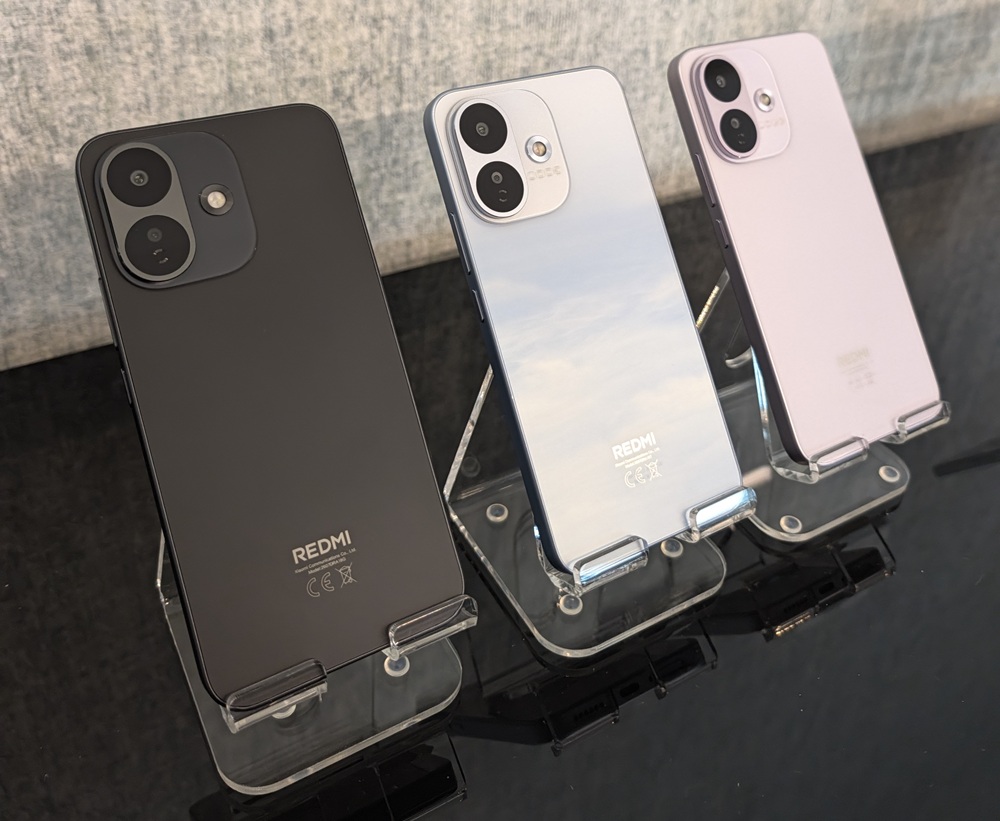 Three Redmi smartphones on display stands viewed from behind, in black, silver, and pink finishes.